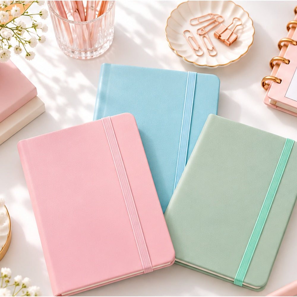 A6 pastel notebooks in pink blue and sage green with elastic bands arranged on a white desk